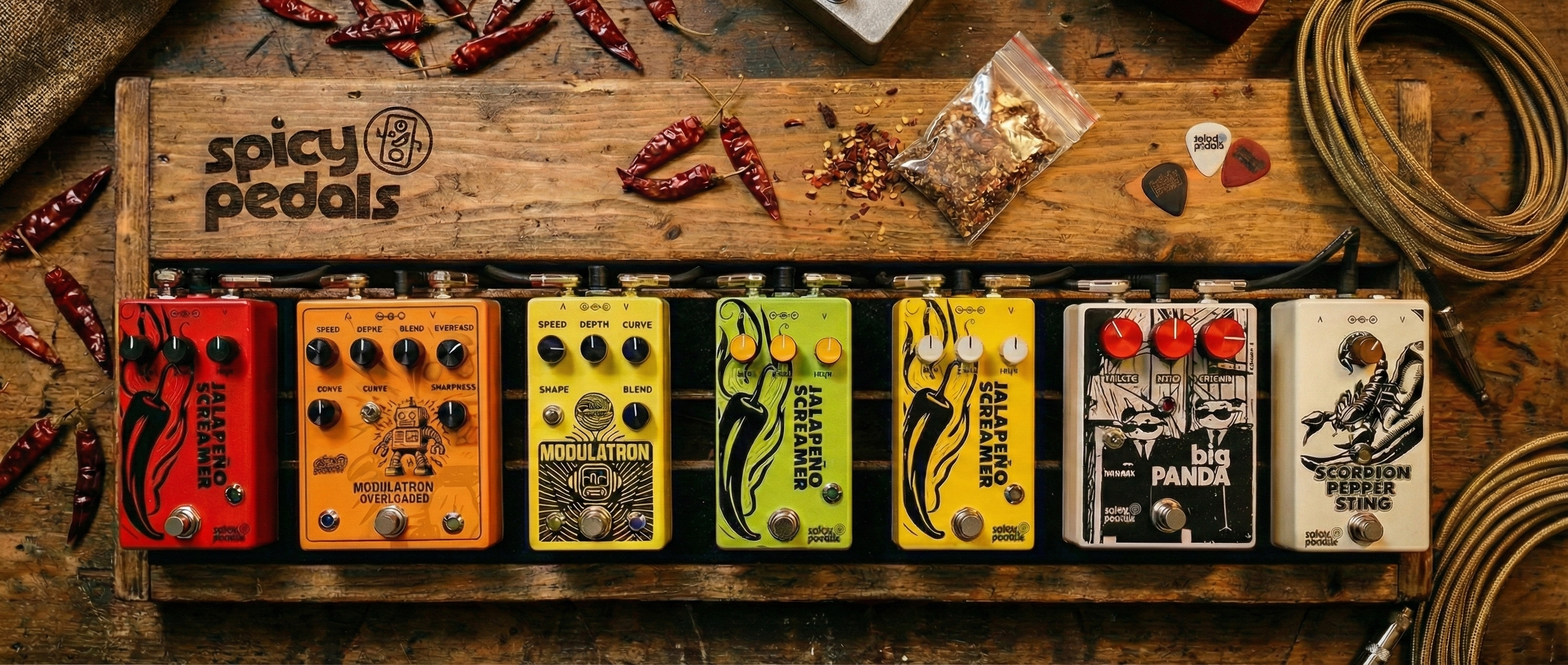 A pedalboard of Spicy Pedals — Jalapeño Screamer, Modulatron, Big Panda and Scorpion Pepper Sting — on a wooden workbench with dried chilies