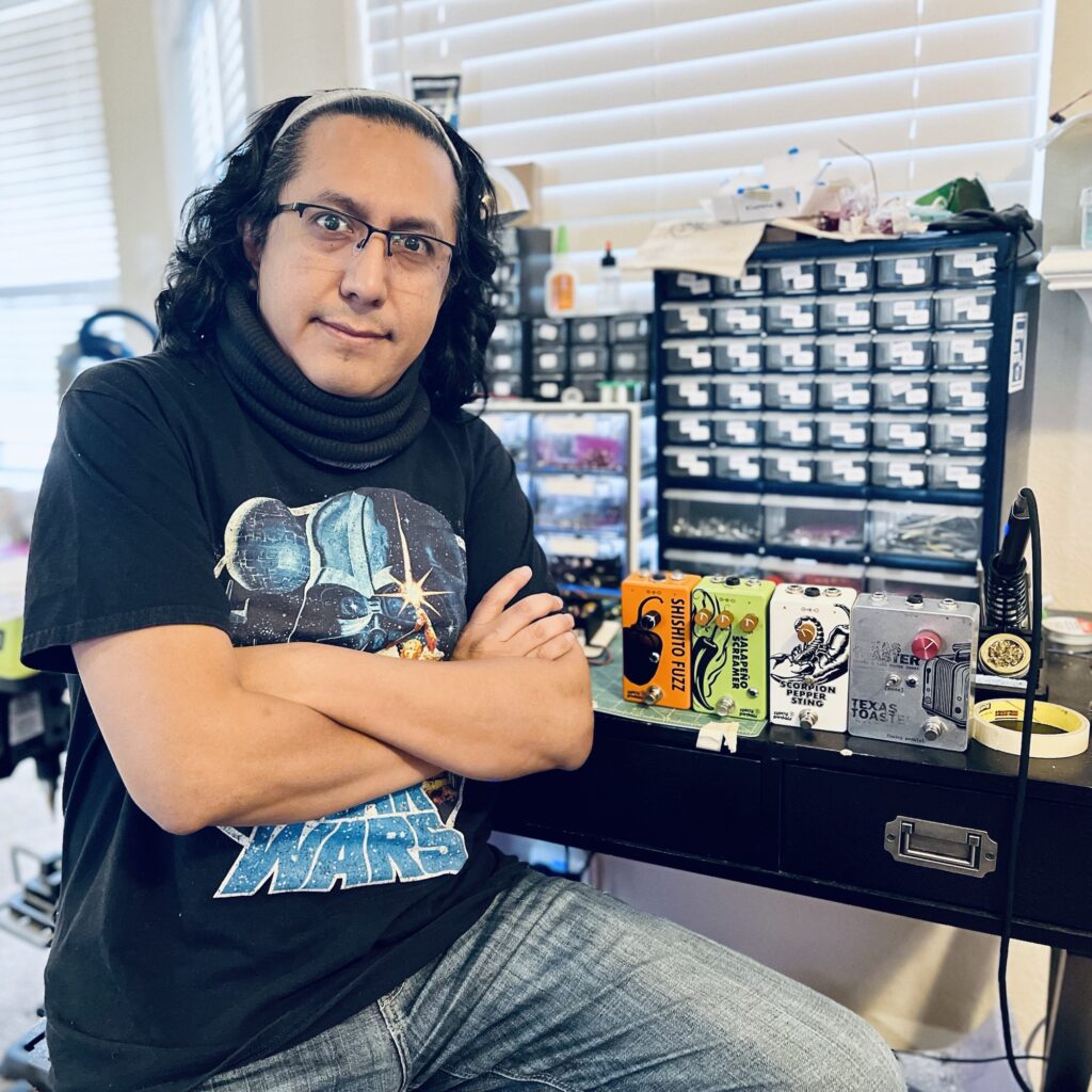 Rolf at his workbench with his arms crossed, in front of a wall of small parts drawers. Four finished pedals stand on the bench beside him — Shishito Fuzz, Jalapeño Screamer, Scorpion Pepper Sting and Texas Toaster — next to a soldering iron on its stand.
