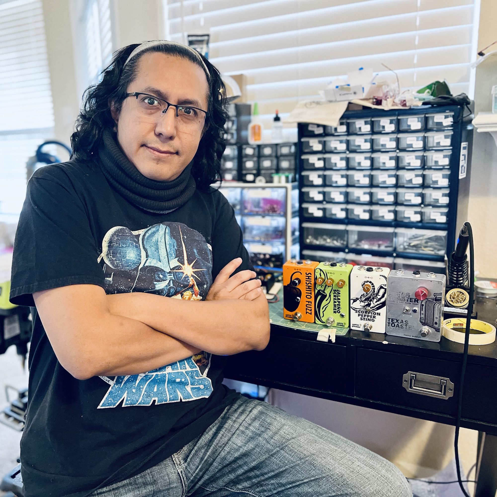 Rolf at his workbench with his arms crossed, in front of a wall of small parts drawers. Four finished pedals stand on the bench beside him — Shishito Fuzz, Jalapeño Screamer, Scorpion Pepper Sting and Texas Toaster — next to a soldering iron on its stand.