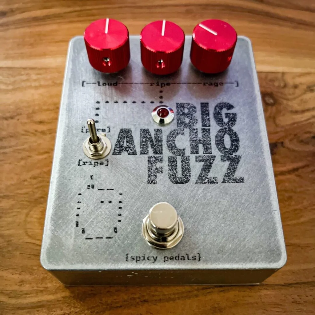 Big Ancho Fuzz on a warm wooden table, seen steeply from above, the three red knobs, the toggle and the {spicy pedals} mark on the face all readable.