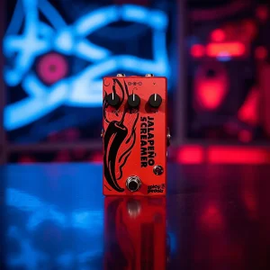 Jalapeño Screamer Rojo on a reflective floor in front of blue and red neon signs, the red enclosure and black knobs picked out against the glow.