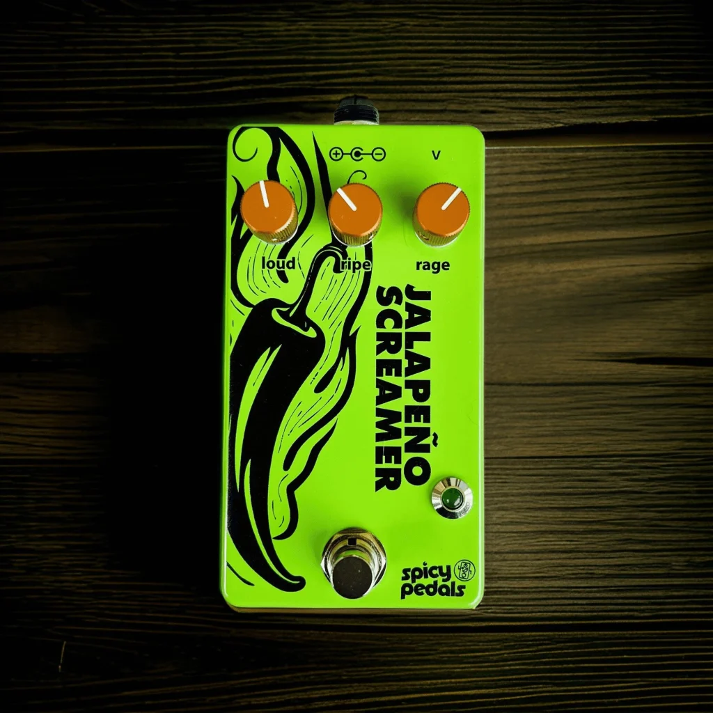 Jalapeño Screamer Verde on dark wooden boards, seen near-frontal at a slight tilt, the orange knobs and the chilli illustration lit from the left.