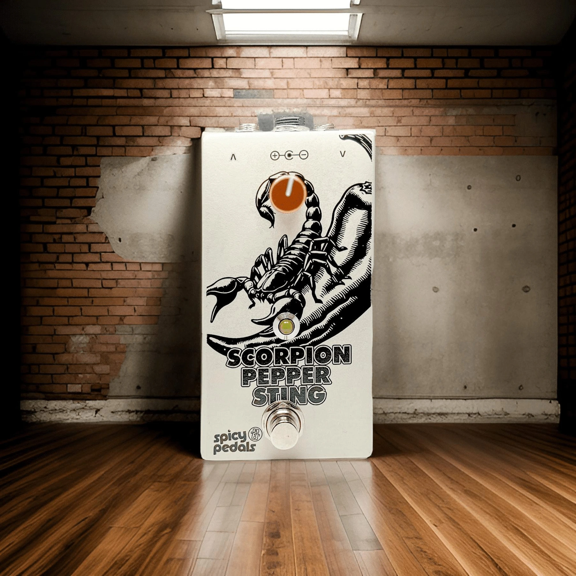 Scorpion Pepper Sting standing against a brick wall on a wooden floor, lit by a fluorescent tube overhead, the scorpion artwork and single orange knob visible.