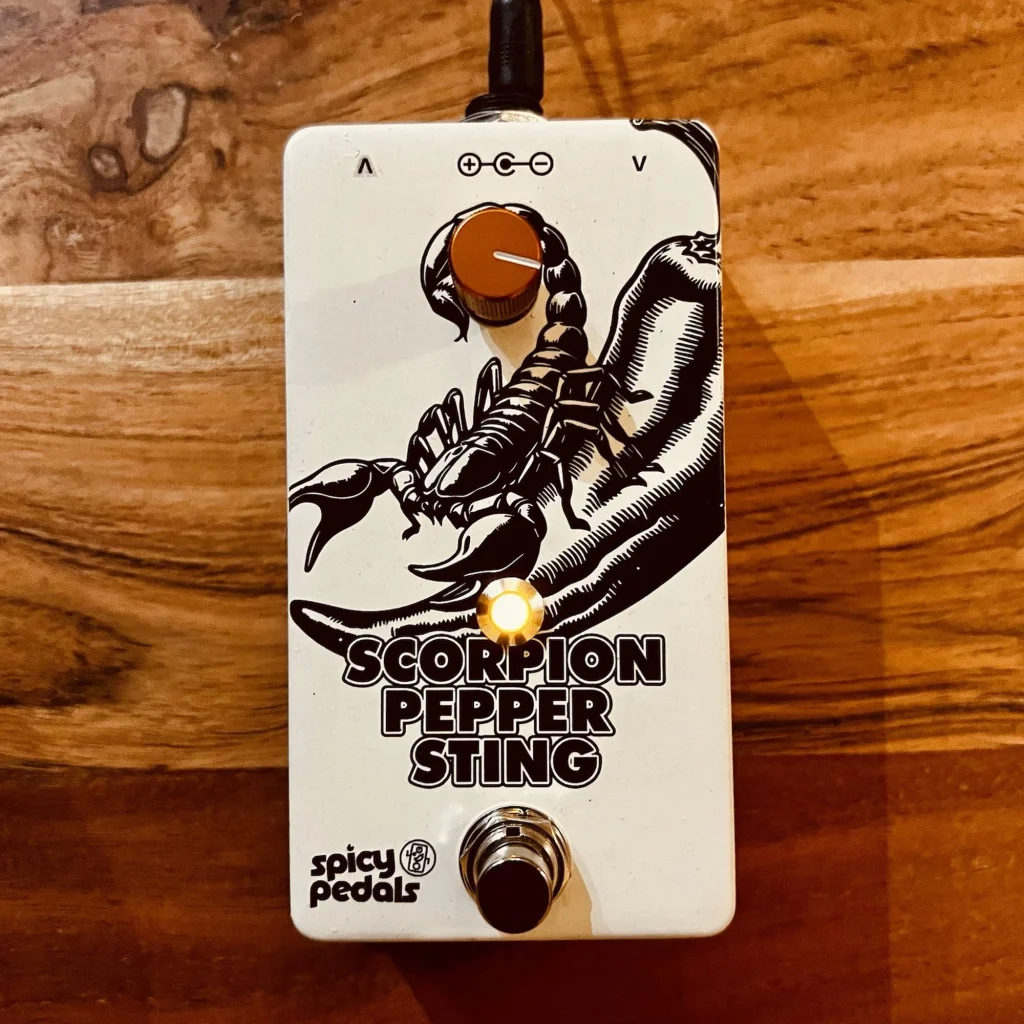 Scorpion Pepper Sting flat on warm wood with a cable plugged into the top, seen from above, the LED glowing amber below the orange knob.