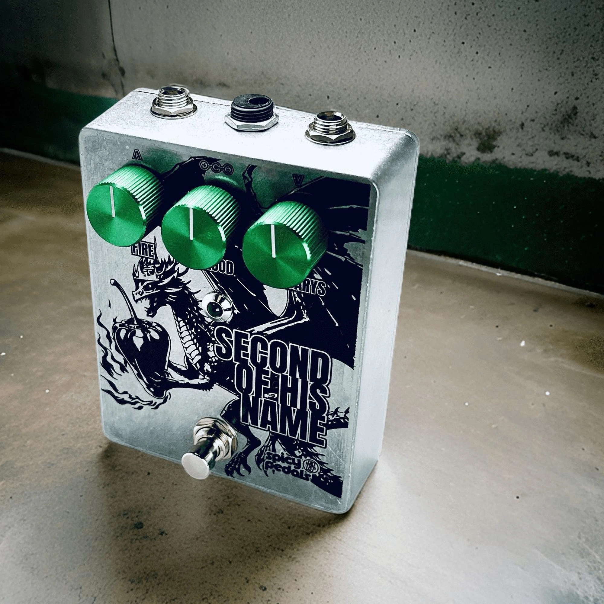 Second Of His Name tilted on a concrete floor against a grey wall, seen from three quarters, the three green knobs and the dragon artwork catching the light.