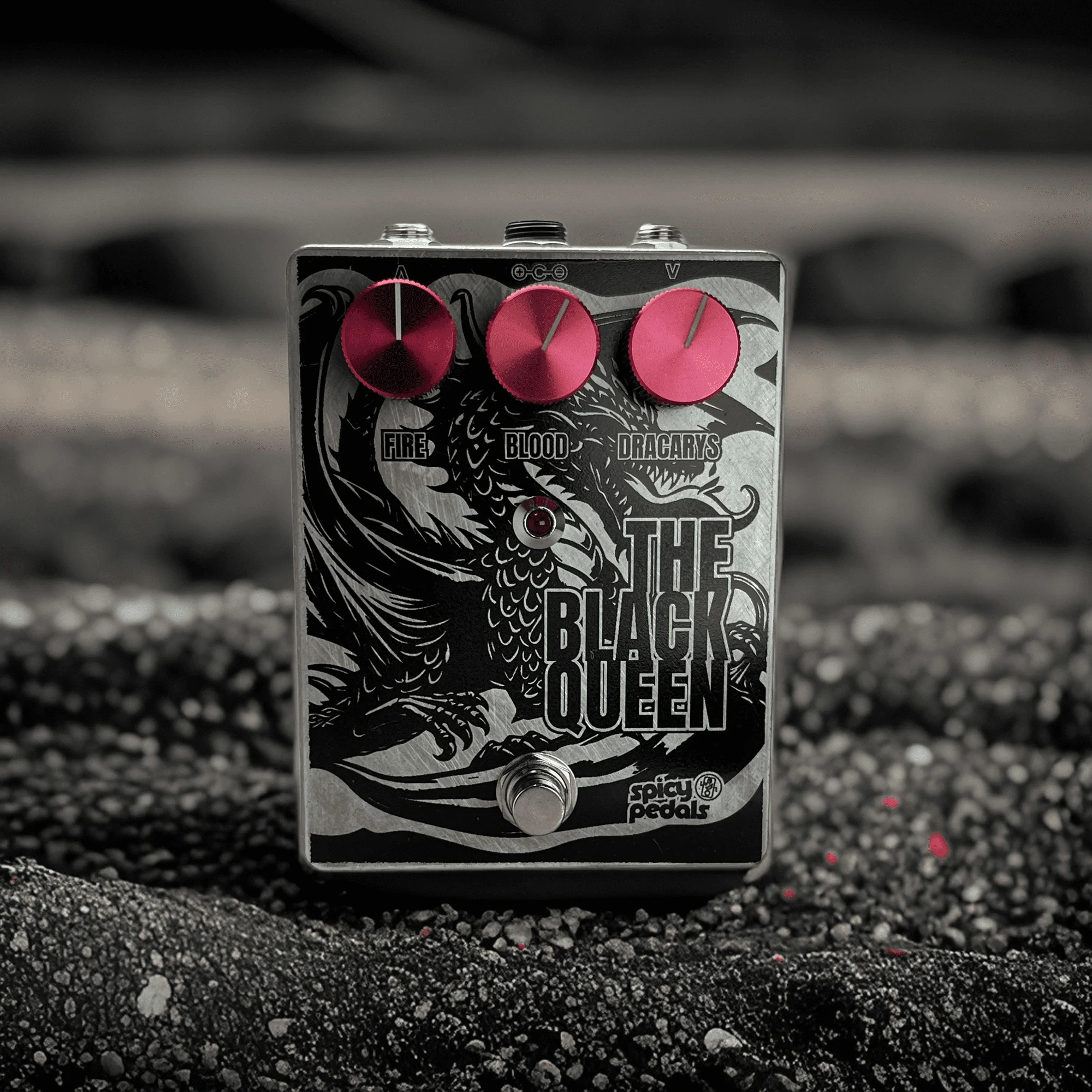 The Black Queen standing on grey gravel in near-monochrome light, seen from the front, the three pink knobs the only colour in the frame.